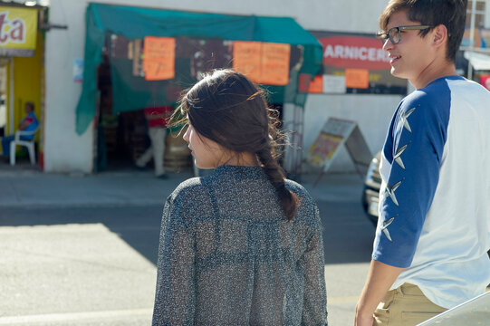 Two people are casually walking on a sunny street, past a store with bright orange awnings and assorted signs.