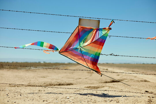 A colorful kite is entangled in a barbed wire fence against a clear blue sky in a barren landscape.
