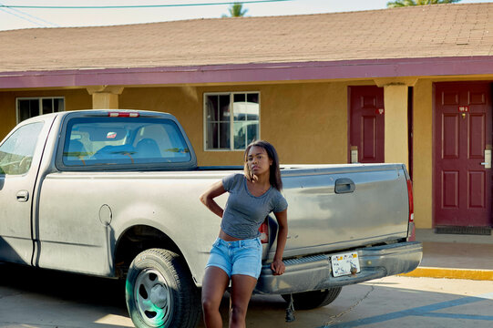 A woman stands in front of a silver pickup truck parked by a mustard-colored building with a red door, her hand on her hip and a contemplative look on her face.