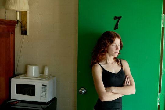 A woman stands in a room with her arms crossed, next to a kitchen counter with a microwave and a coffee maker, looking to the side against a green backdrop.