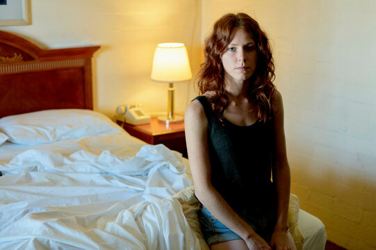 A woman is sitting on the edge of an unmade bed in a room, looking pensively to the side.