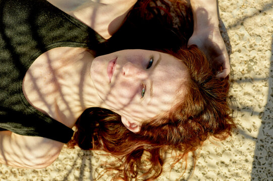 A woman with red hair is lying on a sandy surface, looking up, with sunlight casting shadows across her face.