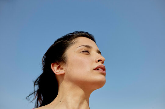 A woman gazes upward against a clear blue sky, showcasing a profile view of her face with her hair swept back.