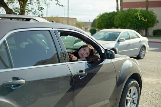 A woman is resting her head on the window frame of a car with her eyes closed, as if she is taking a nap or relaxing.