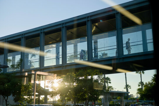 A sunlit modern pedestrian bridge with reflective glass walls, under which the low-hanging sun casts streaks of light and shadow.