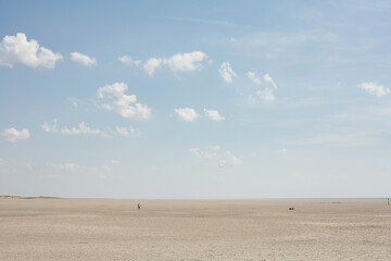 The image showcases a vast, sandy beach with a calm blue sky overhead dotted with white clouds. People appear as tiny figures in the distance, emphasizing the expansive nature of the beach setting.