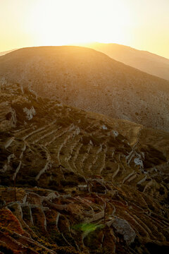 The sun sets behind a hill, casting a warm glow over terraced slopes with a network of paths or steps carved into the landscape.