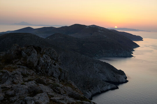 The sun sets behind a mountainous coastline, casting a warm glow over the serene seascape.