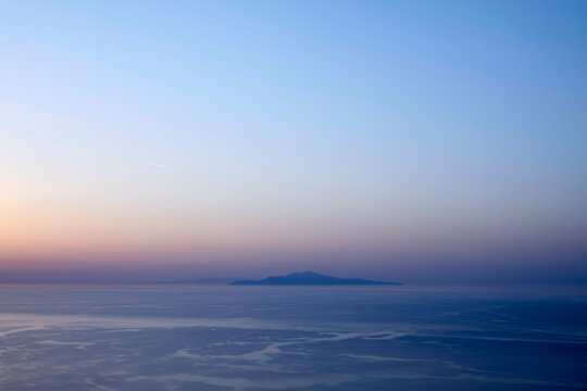 A serene view of a tranquil sea at dusk with a silhouetted island in the distance under a gradient sky transitioning from warm pink to cool blue.