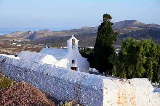 A serene landscape featuring a traditional white-washed church with a bell tower, surrounded by a stone wall, overlooking a hilly terrain with the sea in the distance at dawn.