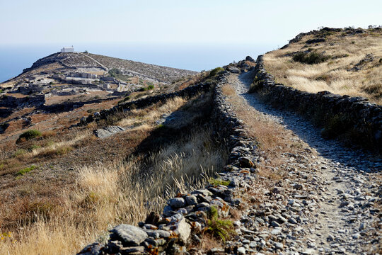 A stone hiking trail leads through a dry, grassy landscape toward a distant overlook with a clear blue sky above.
