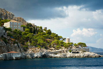 A scenic coastal view featuring a rugged cliffside adorned with Mediterranean-style buildings and lush greenery under a dramatic sky.