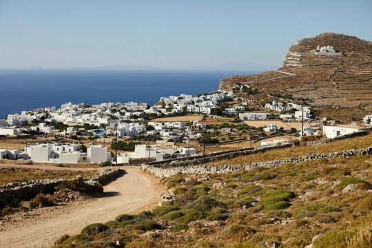 A scenic view of a coastal village with white buildings against a backdrop of clear blue skies, with a dirt path leading through a hilly landscape.