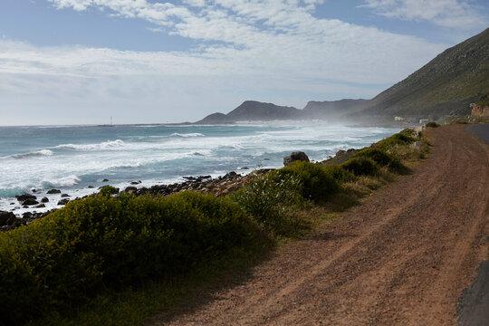 Ocean waves crash against a rocky shoreline parallel to a dirt road, with a mountainous backdrop partially shrouded by mist.