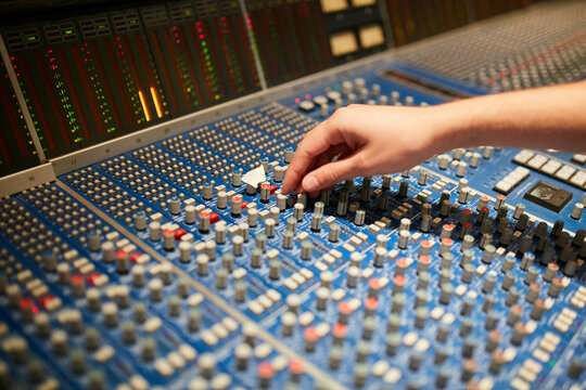 A close-up image of a hand adjusting sliders on a professional audio mixing console with colorful level meters and various knobs.