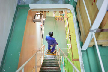 A person in blue coveralls is descending a metal staircase in an industrial environment with yellow safety rails.