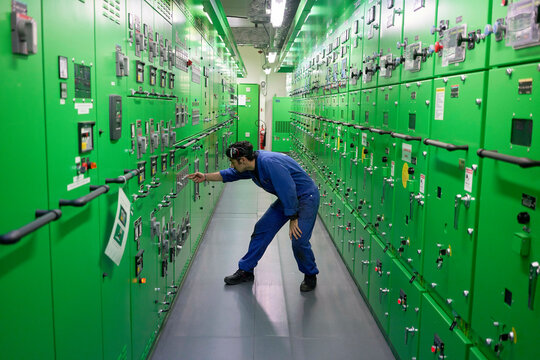 A technician in blue workwear is inspecting or adjusting equipment in a green industrial control room filled with numerous panels and gauges.