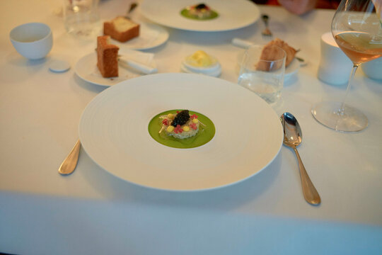A gourmet dish is presented on a large white plate at an elegant dining table with silverware, a glass of wine, and bread on the side.