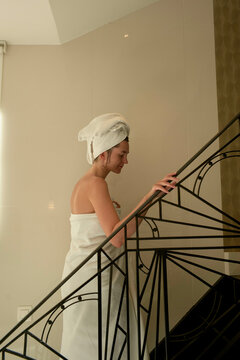 A person wrapped in a towel, with another towel on their head, is ascending a spiral staircase in a bright, modern interior.