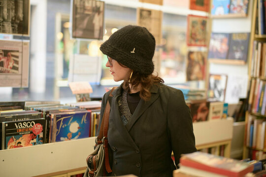 A woman wearing a black hat and coat is browsing a bookshop with a thoughtful demeanor, her attention focused on the books in front of her.