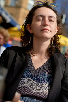 A young adult with closed eyes and a relaxed expression is soaking up the sun, wearing a patterned top, a black jacket, and sunglasses pushed up onto their head.