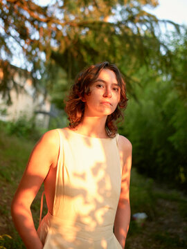 A young woman stands outdoors bathed in the warm glow of sunlight, with the dappled shadow of leaves cast upon her and a backdrop of lush greenery.