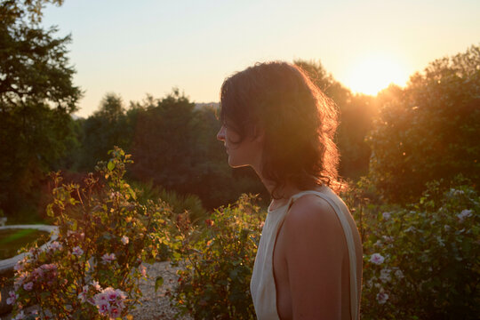 A woman is standing in a garden at sunset, the warm light casting a soft glow on her profile with blooming flowers and greenery in the background.
