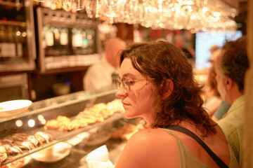 A woman is sitting at a bar with a thoughtful expression, gazing to her side, as the warm ambient lighting illuminates an array of tapas in the background.