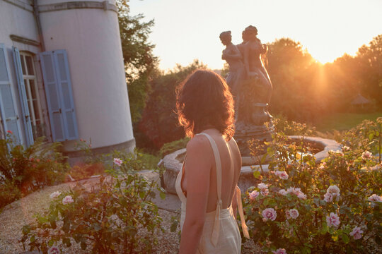 A woman stands in a serene garden at sunset, her silhouette gently outlined by the warm light, with blooming roses and a classic statue in the background.