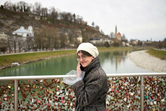 A person stands on a bridge over a river, smiling at the camera, with numerous love locks in the background.