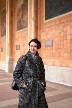 A person is standing in front of an ornate wall with murals, wearing a houndstooth coat and carrying a black bag.