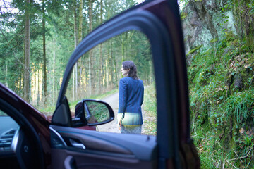 A young woman stands by a forest road, viewed from the open door of a car with the interior partially visible.