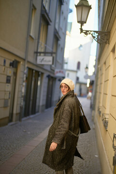 A woman in a fashionable houndstooth coat and white beret looks back over her shoulder while walking down a narrow street with European architecture.