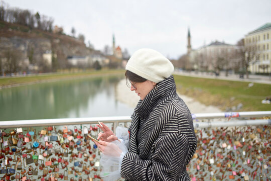 A person stands on a bridge overlooking a river with numerous love locks attached to the railing, while checking their smartphone.