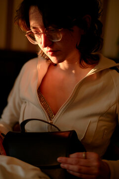 A woman is looking inside a black handbag, illuminated by warm light which highlights her profile and infuses the scene with a soft glow.