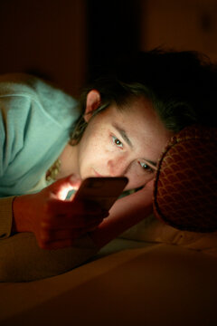 A person is lying in bed, lit by the glow of a smartphone screen in a dark room, their attention captivated by the device.