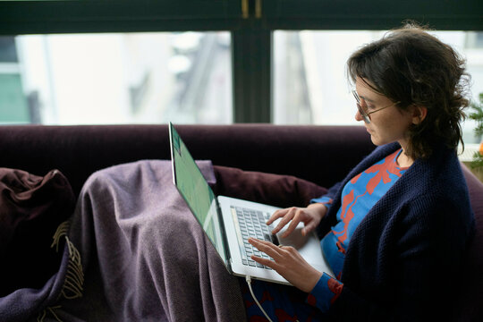 A woman sits comfortably on a purple couch with a blanket, focused on her laptop.