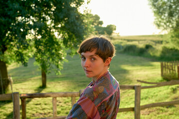 A woman with short hair glances over her shoulder, standing in a field at sunset with a wooden fence in the background.