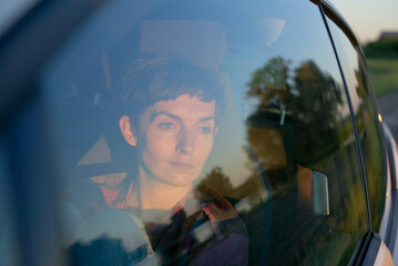 A young woman is gazing out through a car window during twilight, with reflections visible on the glass.
