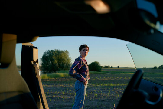 An young woman stands in a field viewed through the open rear window of a car during twilight, with the car interior framing the shot.
