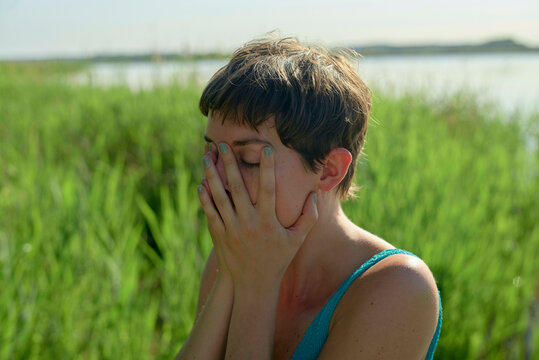 A young woman is sitting outdoors with their eyes covered by their hands, seemingly in a moment of distress or contemplation, with a water body and green reeds in the background.