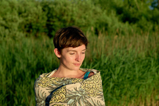A young woman with a pixie haircut is outdoors with eyes closed and a calm expression, bathed in soft sunlight with green foliage in the background.