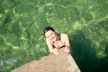 A person is smiling at the camera while holding onto the edge of a pool with clear green water.