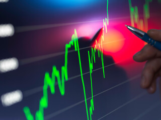 A hand holding a pen points to a bright stock market chart on a digital screen with fluctuating green and red graph lines indicating financial data.