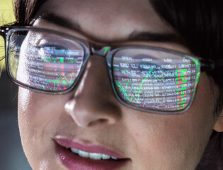 A close-up of a woman's face focusing on her glasses which reflect the colorful glow of a data-driven interface, highlighting numbers and graphs on the screen.