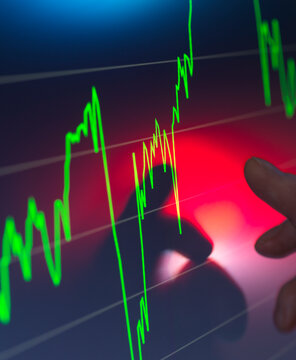 A hand points to a monitor displaying green stock market graphs, with a sharp uptrend, against a red background.