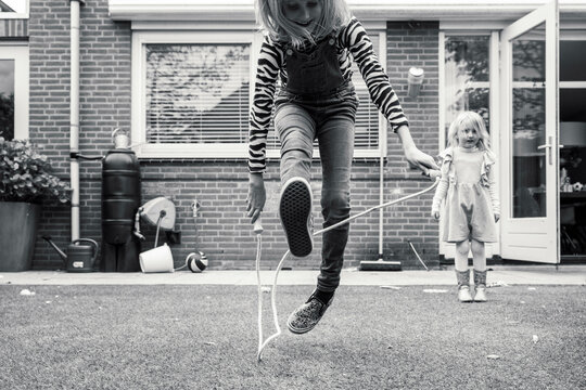 A black and white photo capturing a moment of play, with a girl in mid-air turning a skipping rope and another child watching in the background, set against a residential backdrop.