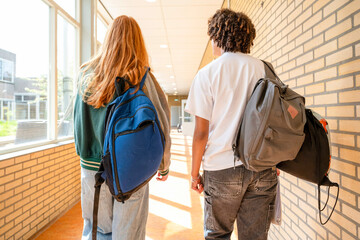 Two students are walking down a well-lit school corridor, seen from the back, carrying backpacks and dressed in casual attire.
