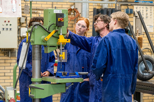 Three people wearing blue work coveralls are attentively observing a green vertical drilling machine in an industrial setting.