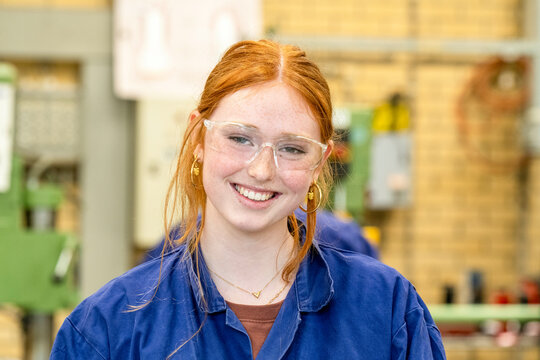 A smiling young woman with red hair, wearing protective glasses and a blue work shirt, is in an industrial or workshop setting.